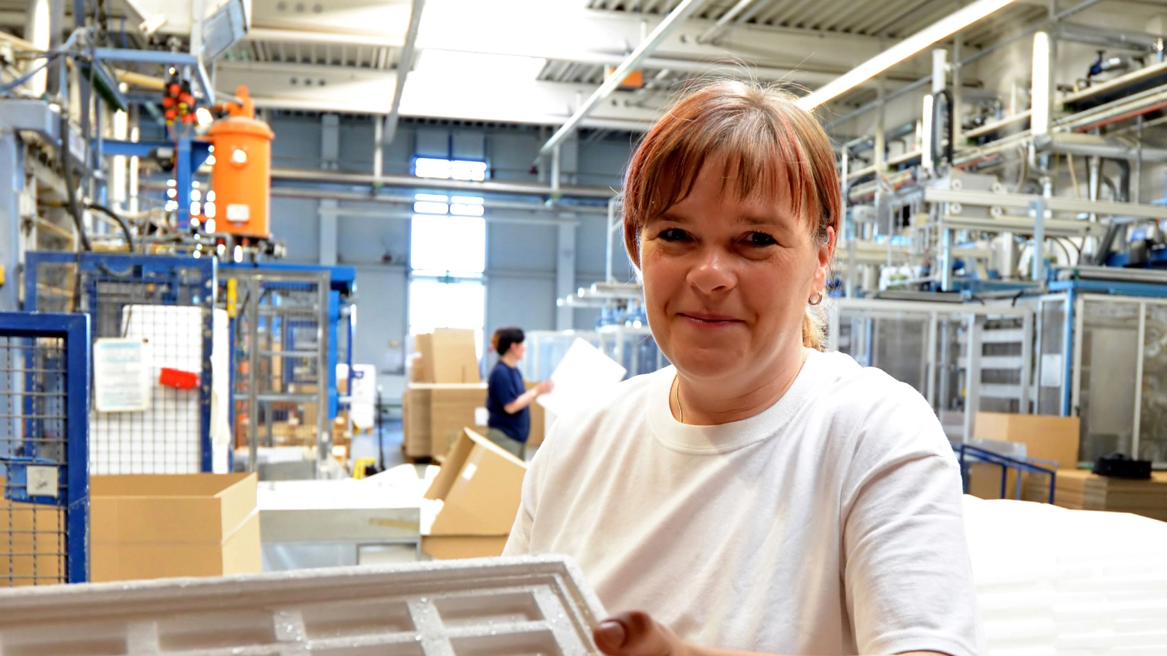 Eine Frau in einer Werkhalle hält eine Styroporkomponente in der Hand und lächelt in die Kamera.