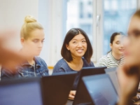 Junge Frau sitzt am Laptop zwischen einigen anderen Seminarteilnehmern und lächelt.