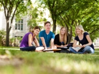 Drei Studentinnen und ein Student sitzen mit Büchern auf einer Wiese und blicken lächelnd in die Kamera.
